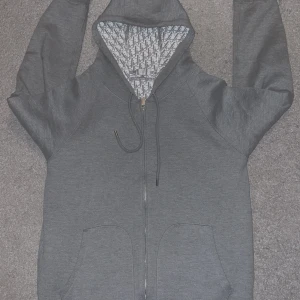 Grå hoodie med mönstrad huva - Säljer en grå hoodie med hel dragkedja och snörning i huvan. Insidan av huvan och hela fodret har ett snyggt grått mönster. Tröjan har broderad på fickan och två stora fickor framtill. Perfekt för dig som gillar exklusiva streetwear-vibes.