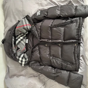 Svart pufferjacka från Burberry - Säljer en svart pufferjacka från Burberry med klassiskt rutigt foder i grått, vitt och rött. Jackan har huva, dragkedja och knappar, samt flera praktiska fickor. Perfekt för kalla dagar och har en modern, oversized look.