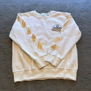 Vit Off-White sweatshirt med print - Vit sweatshirt från Off-White med ikoniskt gult tejp-print på ärmar och rygg samt svart logga framtill. Tröjan har rund hals, ribbade muddar och är tillverkad i mjuk bomull. Perfekt för dig som gillar streetwear och vill sticka ut med en unik design.