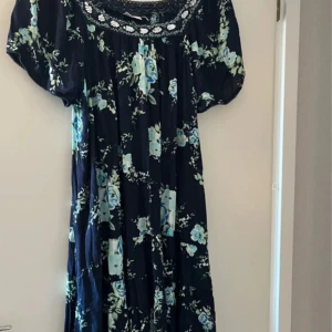Blommig klänning från Isolde - Supersöt långklänning från Isolde med marinblå botten och blommigt mönster i blått/grönt. Klänningen har korta ärmar, volanger och en dekorerad halsringning med paljetter och pärlor. Perfekt för dig som gillar boho-vibe och detaljer.  Köpt på Cassels, i storlek XXL. Den passar upp till 48/50. 100 % viskos, tyget är inte slätt utan lite ”krinklat”. 