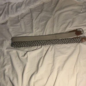 Två flätade bälten - Säljer två stiliga flätade bälten. Ett i beige och ett i grått med bruna läderdetaljer. Perfekta för att ge en avslappnad men ändå snygg look. Bältena har en elastisk design som gör dem bekväma att bära.