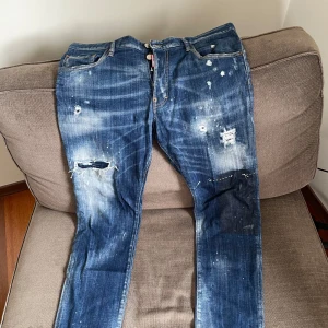 Blå jeans från Dsquared2 - Säljer ett par blå jeans från Dsquared2 med slitna detaljer och färgstänk för en edgy look. Jeansen har en klassisk femficksdesign och knappgylf. Perfekta för dig som vill ha en unik stil.
