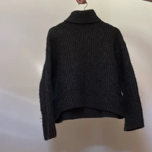 Mörkgrå stickad polotröja - Säljer en mysig mörkgrå stickad polotröja med hög krage. Tröjan har en avslappnad passform och långa ärmar, perfekt för kyliga dagar. Den ribbade strukturen ger en stilren look.