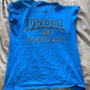 Blå t-shirt med text - Snygg blå t-shirt med texten 'London England' i glittriga stenar. Perfekt för en avslappnad stil med korta ärmar och rund hals. Passar bra till jeans eller shorts för en cool look. Några stenar har gått bort! 