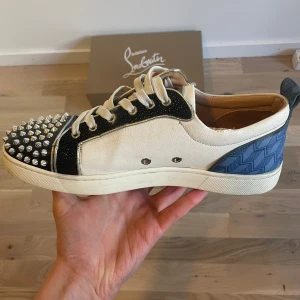 Christian Louboutin - Snygga sneakers från Christian Louboutin med en unik design. Skorna har en vit bas med blå och svarta detaljer samt silverfärgade nitar på tån. Den ikoniska röda sulan ger en lyxig touch. Perfekta för den som vill sticka ut med stil.