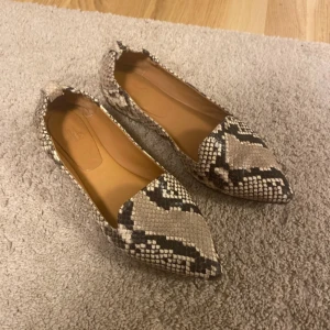 Ormskinnsmönstrade ballerinaskor - Snygga ballerinaskor med ormskinnsmönster och spetsig tå. Perfekta för att ge en trendig touch till din outfit. Skorna har en elegant design och är i en neutral färgskala som passar till det mesta.