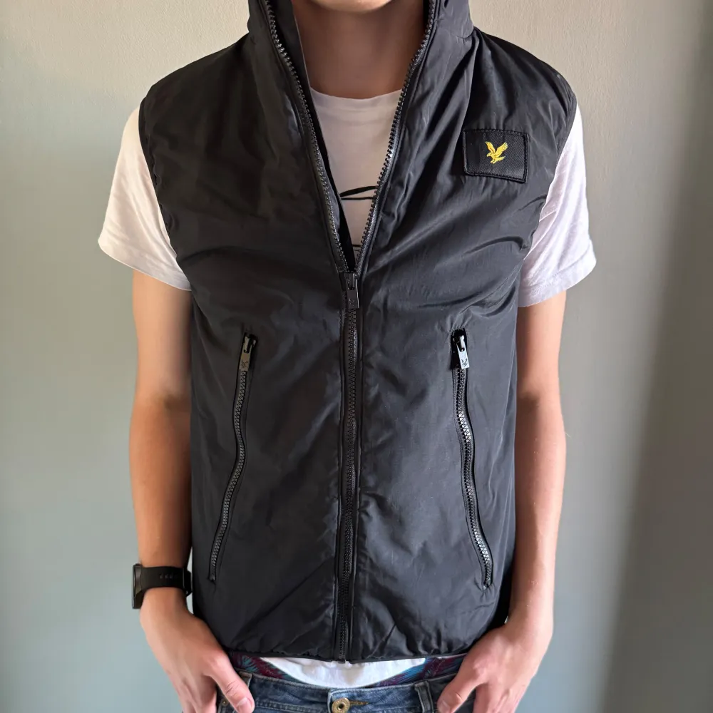 Snygg svart väst från Lyle & Scott med dragkedja och två praktiska fickor framtill. Västen har en stilren design med en liten logotyp på bröstet. Perfekt för lager-på-lager under kyligare dagar. Säljer pga är för liten för mig . Takit.