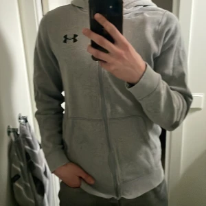 Grå hoodie från Under Armour - Säljer en grå hoodie från Under Armour med dragkedja och huva. Perfekt för en avslappnad stil. Den har en lös passform och är tillverkad i ett mjukt material. Storlek S/M