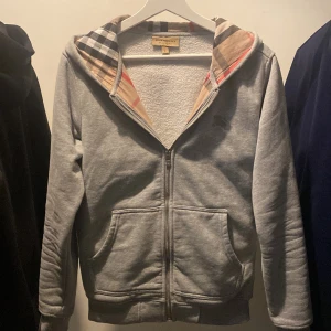 Grå hoodie från Burberry - Säljer en stilren grå hoodie från Burberry med dragkedja och klassiskt rutigt mönster i luvan. Tröjan har broderad logga på ärmen och praktiska fickor framtill. Perfekt för en avslappnad och trendig look.