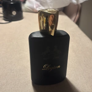 Legion parfym från Fragrance World - Elegant parfymflaska i svart med guldlock från Fragrance World. Flaskan har en stilren design med namnet 'Legion' i guldtext. Locket är triangulärt med en dekorativ gravyr på toppen.