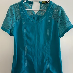 Turkos blus med spetsdetaljer - Snygg turkos blus med korta spetsärmar och v-ringning. Blusen har en elegant spetsdetalj på axlarna och övre delen av ryggen. Perfekt för en stilren look.