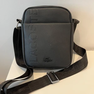Svart axelväska från Lacoste - Snygg svart axelväska från Lacoste med justerbar axelrem och dragkedja. Väskan har en stilren design med Lacoste-loggan präglad på framsidan. Perfekt för att bära dina dagliga nödvändigheter med stil.