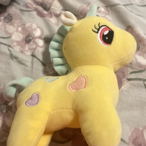 Gul enhörningsmjukisdjur med hjärtan - Söt gul enhörning i mjukt tyg med pastellfärgade hjärtan på kroppen. Den har en mintgrön man och svans, samt stora broderade ögon. Perfekt som kramdjur eller dekoration i barnrummet.