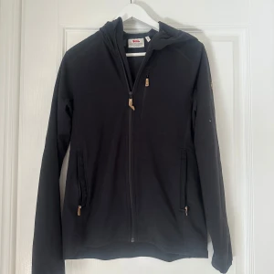 Svart fleecejacka från Fjällräven - Säljer en svart fleecejacka från Fjällräven med dragkedja och huva. Jackan har långa ärmar och praktiska fickor med dragkedja. Perfekt för kyliga dagar.