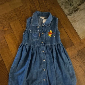 Blå jeansklänning med Nalle Puh-motiv - Charmig ärmlös jeansklänning från Disney med ett broderat Nalle Puh-motiv på bröstet. Klänningen har knappar framtill och en liten bröstficka med texten 'Winnie the Pooh'. Perfekt för små äventyrare!