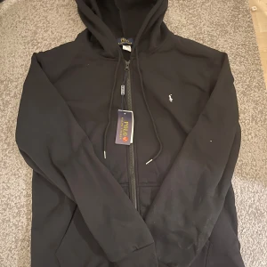 Svart zip hoodie från Ralph Lauren (Storlek L)  - Säljer en stilren svart hoodie från Ralph Lauren med dragkedja och broderad logga på bröstet. Perfekt för en avslappnad look. Storlek L.