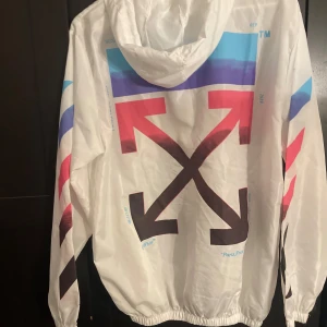 Vit vindjacka från Off-White - Säljer en vit vindjacka från Off-White med färgglada detaljer och ikoniska pilar på ryggen. Jackan har dragkedja framtill och långa ärmar. Perfekt för en stilren look med en touch av färg.