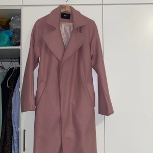 Rosa kappa från Bik Bok - Säljer en stilren rosa kappa från Bik Bok i storlek L. Den har en klassisk krage och långa ärmar, perfekt för kyligare dagar. Kappan har en elegant design med dolda fickor på sidorna.