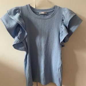 Blå ribbad topp från Zara - Säljer en ljusblå ribbad topp från Zara med volangdetaljer på ärmarna. Toppen har en rund halsringning och korta ärmar som ger en unik look. Perfekt för att piffa upp garderoben!