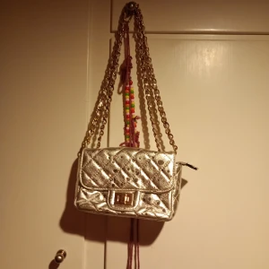 Silverfärgad quiltad axelväska - Snygg guldig axelväska med quiltat mönster och kedjeaxelrem. Perfekt för att ge en glittrig touch till din outfit. Väskan har en praktisk dragkedja och ett elegant spänne framtill.