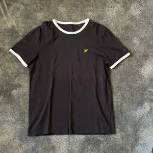 Svart Lyle & Scott t-shirt  - Snygg svart t-shirt från Lyle & Scott med vit kant vid hals och ärmslut. Den har en liten gul logga på bröstet. Perfekt för en stilren look!