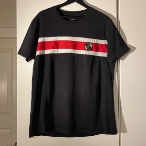 Svart t-shirt med röd och vit rand från Glorious Gangsta - Snygg svart t-shirt från Glorious Gangsta med en bred röd och vit rand över bröstet. T-shirten har en kortärmad design och ett märke på bröstet. Perfekt för en stilren look.