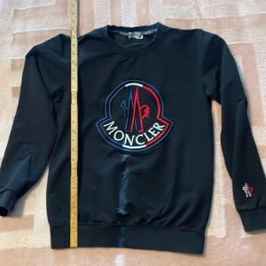 Svart sweatshirt från Moncler - Säljer en svart sweatshirt från Moncler med ett stort broderat logotypmotiv på framsidan. Tröjan har långa ärmar och är tillverkad i bomull med en liten del lycra för extra komfort. Perfekt för en stilren och avslappnad look.