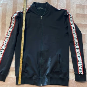 Svart jacka från Givenchy - Säljer en stilren svart bomberjacka från Givenchy med dragkedja framtill. Jackan har långa ärmar med Givenchy-logga längs sidorna i vitt och rött. Perfekt för en trendig look! Lite småfleckar. Kolla bild. 