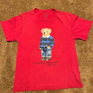 Röd t-shirt med Polo Bear från Ralph Lauren - Säljer en röd t-shirt från Ralph Lauren med en ikonisk Polo Bear-tryck på framsidan. Björnen har en blå och svart randig tröja och jeans. Perfekt för en avslappnad stil med en touch av lyx.
