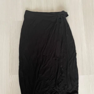 Svart omlottkjol  - Snygg svart omlottkjol från Pine Ridge i mjukt material. Kjolen har en knytning i midjan som ger en fin passform. Perfekt för en stilren look.