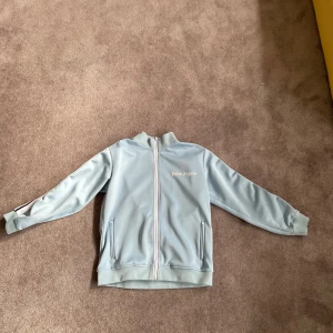 Ljusblå jacka från Palm Angels - Säljer en stilren ljusblå jacka från Palm Angels med dragkedja framtill. Jackan har långa ärmar och ribbade muddar vid ärmslut och nederkant. Perfekt för en avslappnad look.