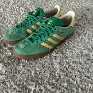 Adidas gazelle - Snygga gröna Adidas gazelle sneakers med klassiska gula ränder och brun sula. Skorna har snörning och är tillverkade i mocka för en stilren look. Perfekta för en sportig stil.