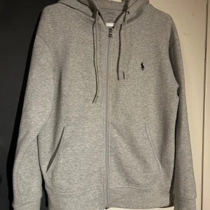 Grå hoodie från Polo Ralph Lauren - Säljer en stilren grå hoodie från Polo Ralph Lauren med dragkedja och justerbar huva. Den har en broderad logga på bröstet och praktiska fickor framtill. Perfekt för en avslappnad look.