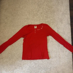 Röd långärmad topp med text - Säljer en röd långärmad topp från The T-shirt Store med texten 'love forever' broderad vid halsen. Toppen har knappar framtill och är tillverkad i ett mjukt material. Perfekt för en avslappnad stil.