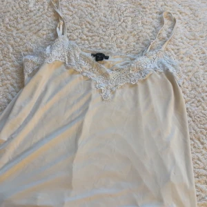 Beige linne med spetsdetaljer - Säljer ett elegant beige linne med smala axelband och vacker spetsdetalj vid halsringningen. Perfekt för en stilren och feminin look.
