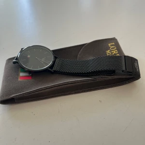 Svart klocka från Lorenzo Milano - Stilren svart klocka från Lorenzo Milano med en minimalistisk urtavla och italiensk flagga. Klockan har ett svart mesh-armband som ger en modern look. Perfekt för dig som gillar enkel och elegant design.