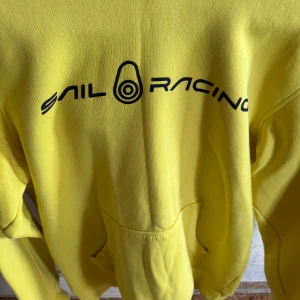 Gul hoodie från Sail Racing - Säljer en snygg gul hoodie från Sail Racing med svart logga på bröstet och en praktisk magficka. Den har en justerbar huva med svarta snören och ribbade muddar vid ärmslut. Perfekt för en sportig look!