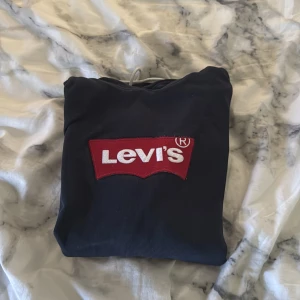 Marin blå hoodie från Levi's - Säljer en svart hoodie från Levi's med det klassiska röda och vita logotypen på bröstet. Tröjan har en justerbar huva med vita snören och en praktisk magficka. Perfekt för en avslappnad stil.