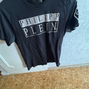 Svart t-shirt från Philipp Plein - Snygg svart t-shirt från Philipp Plein med glittrande logotyp framtill och PP-märke på ärmen. Perfekt för dig som vill ha en stilren men ändå iögonfallande look. Tillverkad i mjukt material för extra komfort. S/M