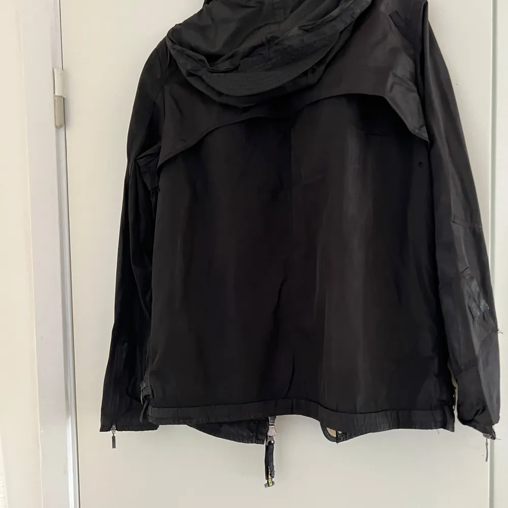 Säljer en svart vindjacka från Parajumpers med dragkedja och flera praktiska fickor. Jackan har en justerbar huva och detaljer som spännen och logotyp på ärmen. Perfekt för blåsiga dagar.. Takit.