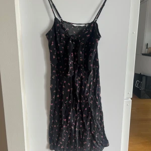 Svart blommig klänning från & Other Stories - Superfin svart sommarklänning från Urban Outfitters med små rosa blommor och gröna blad. Klänningen har smala axelband och en knytning framtill.