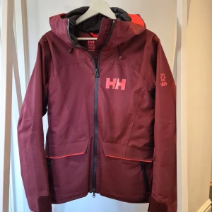Helly Hansen alpinjacka strl M - Skidjacka från Helly Hansen. Vattentät, andas och lätt fodrad med primaloft.  Praktiska fickor med dragkedjor fram och en innerficka med dragkedja och en nätficka. Liftkortsficka på vänster ärm. Huvan går att ha över hjälmen. Snölås. Endast använd för alpin skidåkning  Recco. 