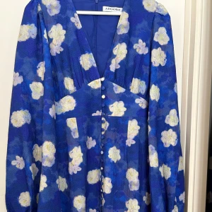 ADOORE Positano - Snygg blå klänning från ADOORE med blommigt mönster i ljusa toner. Klänningen har långa ärmar och knappar framtill som ger en elegant touch. Perfekt för en stilfull look.