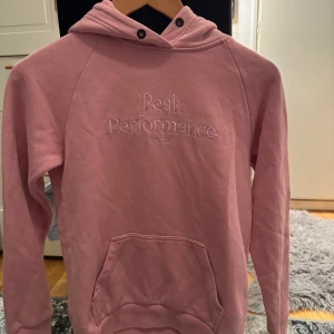 Rosa hoodie från Peak Performance - Säljer en snygg rosa hoodie från Peak Performance med broderad logga på bröstet. Tröjan har en klassisk känguruficka och en bekväm huva. Perfekt för en avslappnad stil.