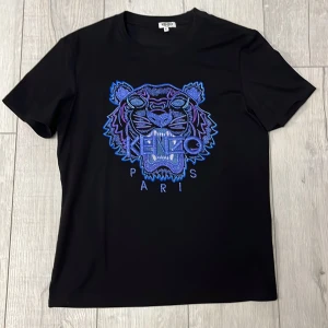 Svart t-shirt med tigerbroderi från Kenzo - Snygg svart t-shirt från Kenzo med ett stort, färgglatt tigerbroderi i blått och lila på framsidan. Perfekt för att ge din outfit en cool touch. T-shirten har en normal passform och korta ärmar.