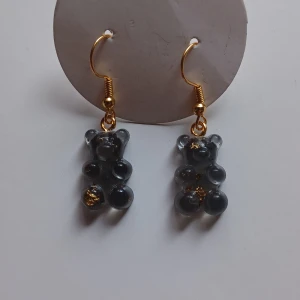 Guldiga örhängen med björndetaljer - 2a utrensning! 4 olika par, säg till vilket par du vill ha vid köp. Snygga örhängen med guldiga krokar och björndetaljer i en mörk nyans. Perfekta för att ge en lekfull touch till din stil. De små björnarna har en glittrig effekt som fångar ljuset fint.