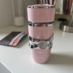 Yara parfym från Lattafa - Elegant parfymflaska i ljusrosa med silverdetaljer från Lattafa. Flaskan har en unik design med dekorativa band och en rund emblem på framsidan. Perfekt för den som söker en stilfull och modern doftupplevelse. Använd 2-3 gånger 