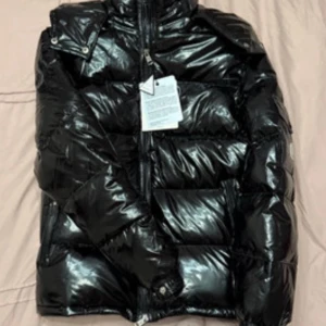 Svart dunjacka från Moncler - Säljer en snygg svart dunjacka från Moncler. Jackan har en glansig finish och är utrustad med dragkedja framtill. Perfekt för kyliga dagar när du vill hålla stilen. 🖤 med pris lapp på ny 
