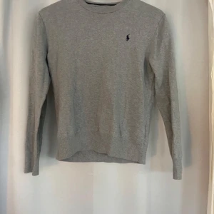 Grå tröja från Polo Ralph Lauren - Säljer en stilren grå stickad tröja från Polo Ralph Lauren. Tröjan har en diskret logga på bröstet och är tillverkad i ett mjukt material. Perfekt för en avslappnad look.
