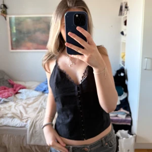 Svart korsettopp med spetsdetaljer - Snygg svart korsettopp med spetsdetaljer längs kanterna och knäppning framtill. Perfekt för en stilren look.